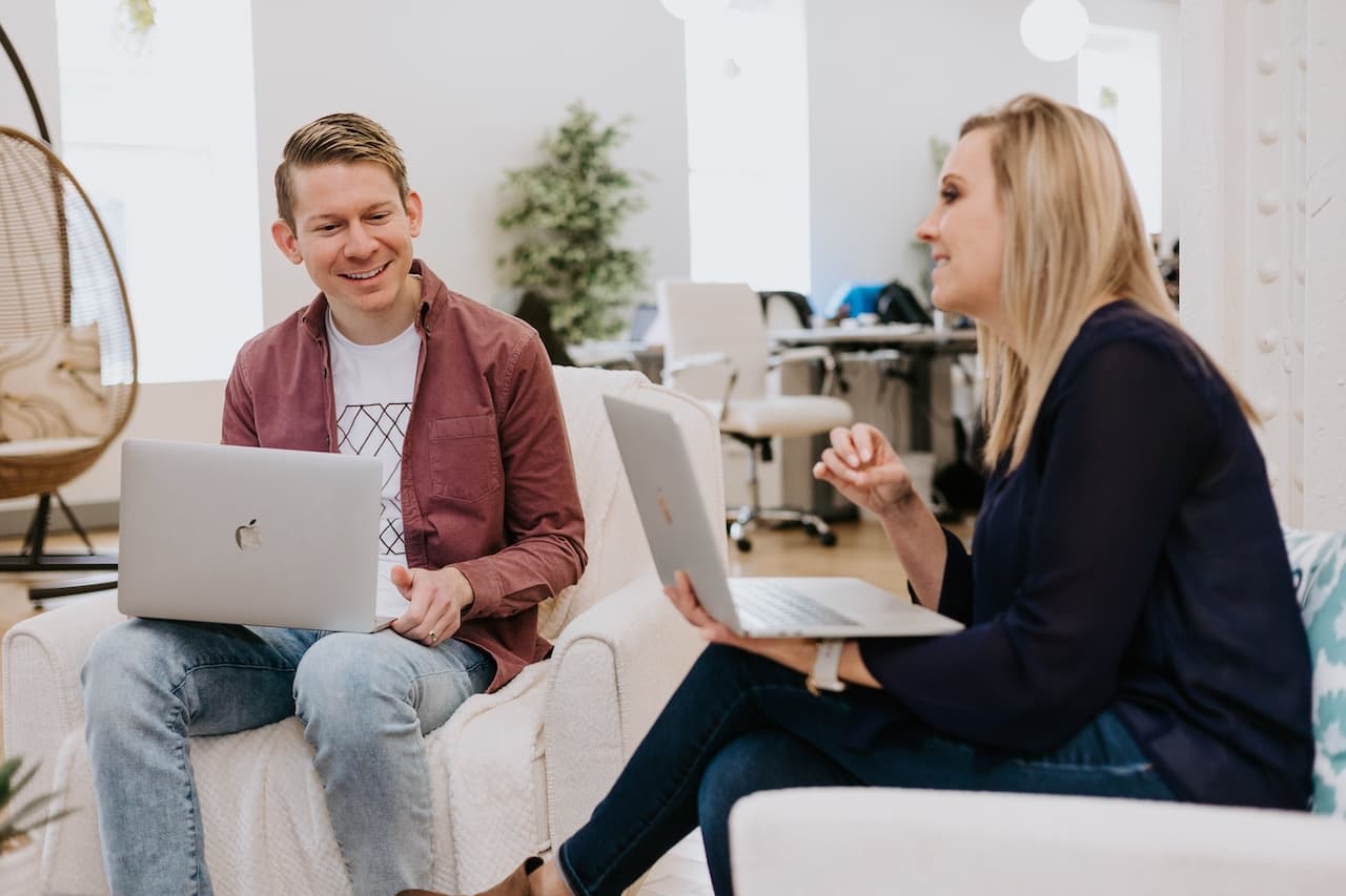 Two colleagues having a conversation while working on laptops in an office