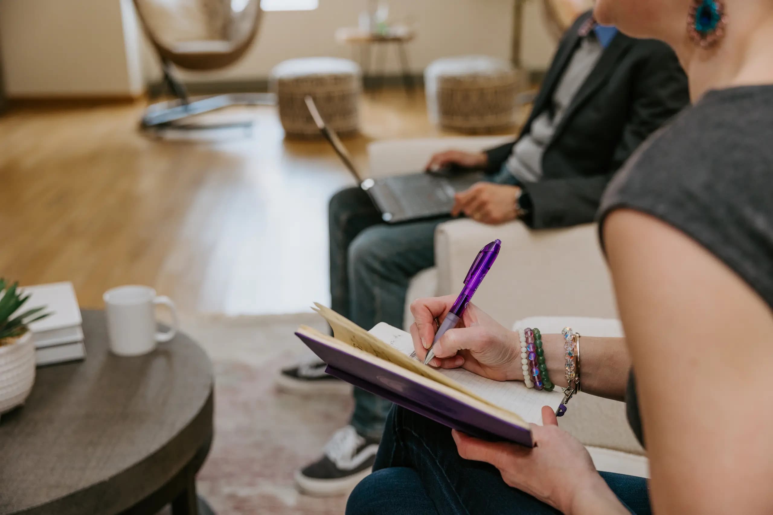 Close-up of a woman's hands writing in a notebook with a purple pen during a meeting. She is wearing colorful bracelets, seated next to a man using a laptop in a modern, well-lit office lounge with comfortable seating.