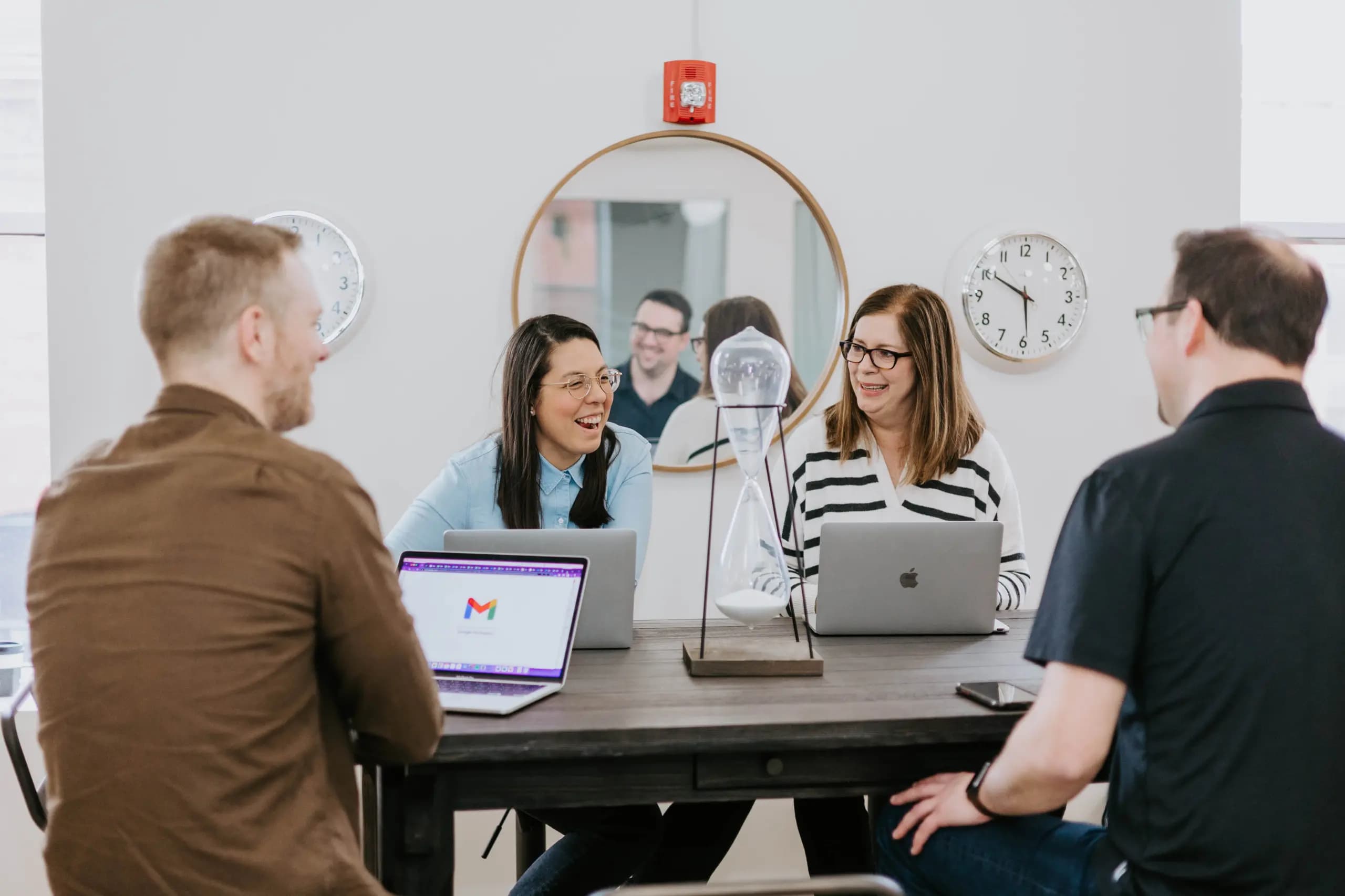 Four colleagues engaging in a discussion at a long table, with laptops open in front of them. The setting includes a large round mirror reflecting the group, and wall clocks showing different times, creating a modern office atmosphere.