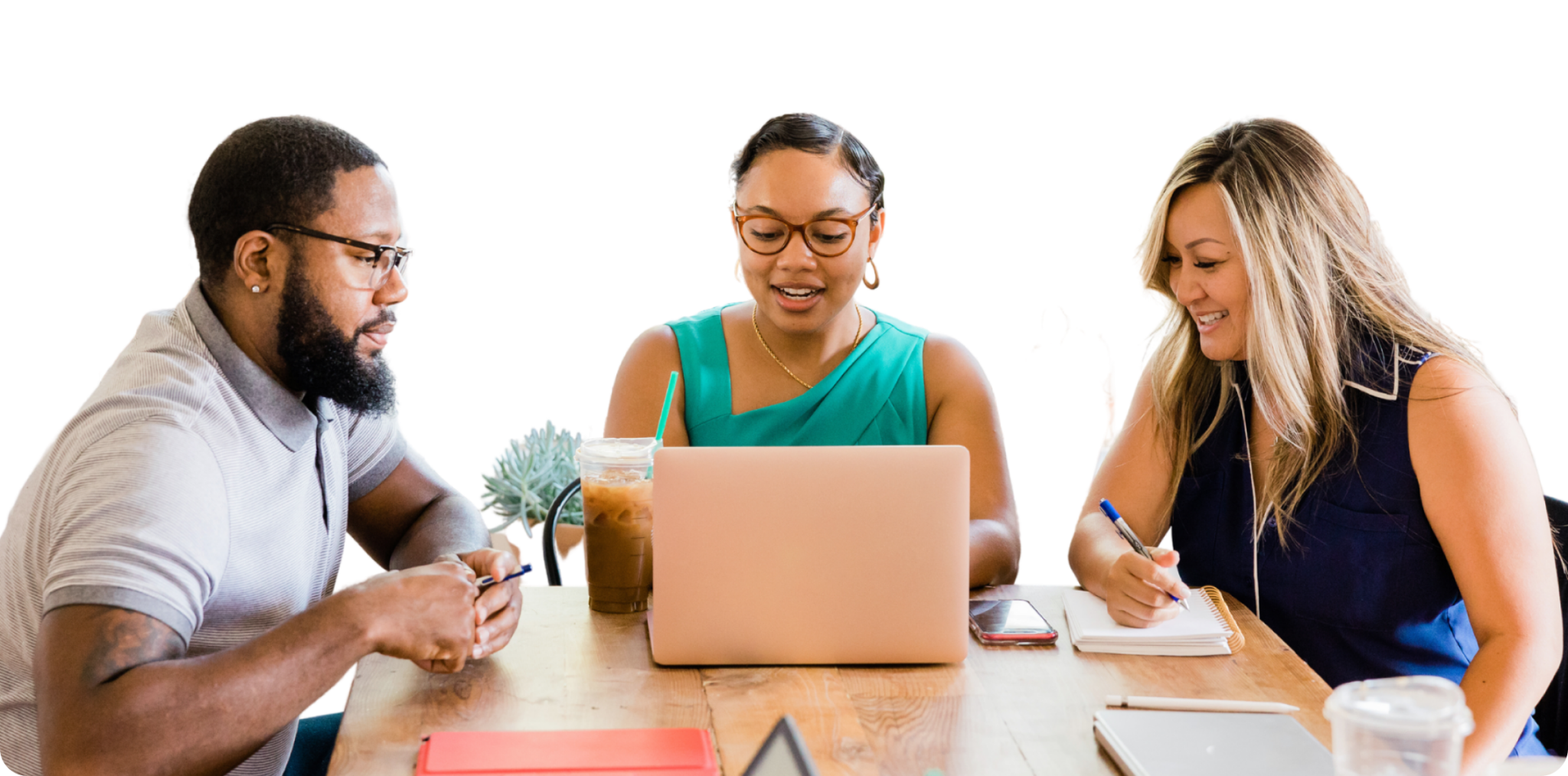 Three people are gathered around a wooden table. They are having a meeting. One person is using a laptop, one is taking notes with a pen and paper, and the third is looking at the laptop screen. Multiple items, including notebooks and drinks, are on the table.